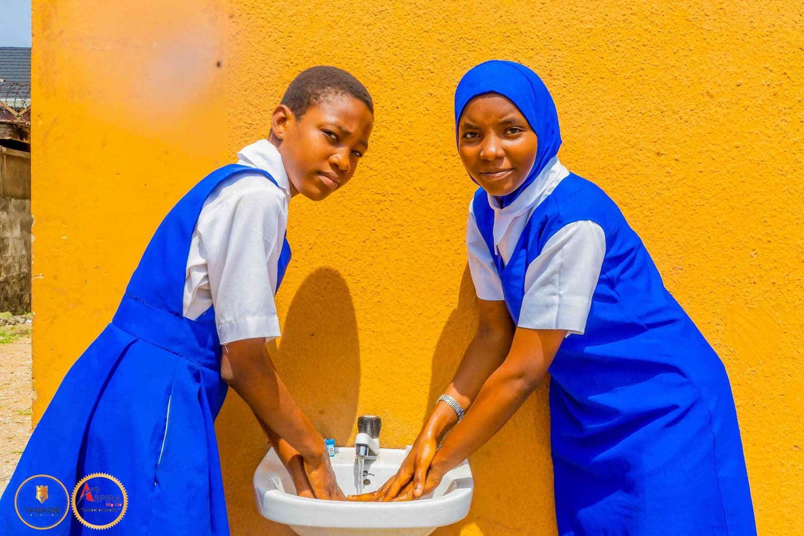 Two students wash their hands together at a basin against a bright yellow wall